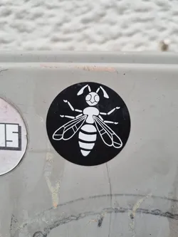 A black and white round sticker featuring a stylized wasp illustration. The wasp is white against a black circular background. The sticker appears to be affixed to a light grey surface outdoors.