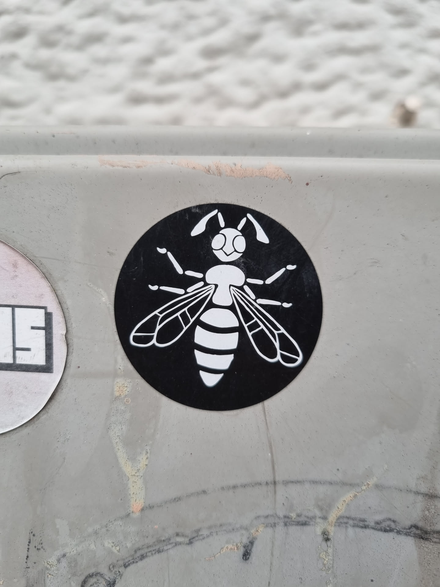 A black and white round sticker featuring a stylized wasp illustration. The wasp is white against a black circular background. The sticker appears to be affixed to a light grey surface outdoors.