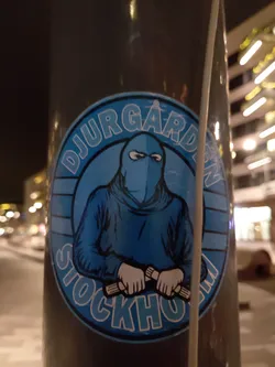 A blue and white circular sticker featuring a hooded figure holding what appears to be two batons or similar objects. The text "DJURGÅRDEN STOCKHOLM" is prominently displayed around the central image.