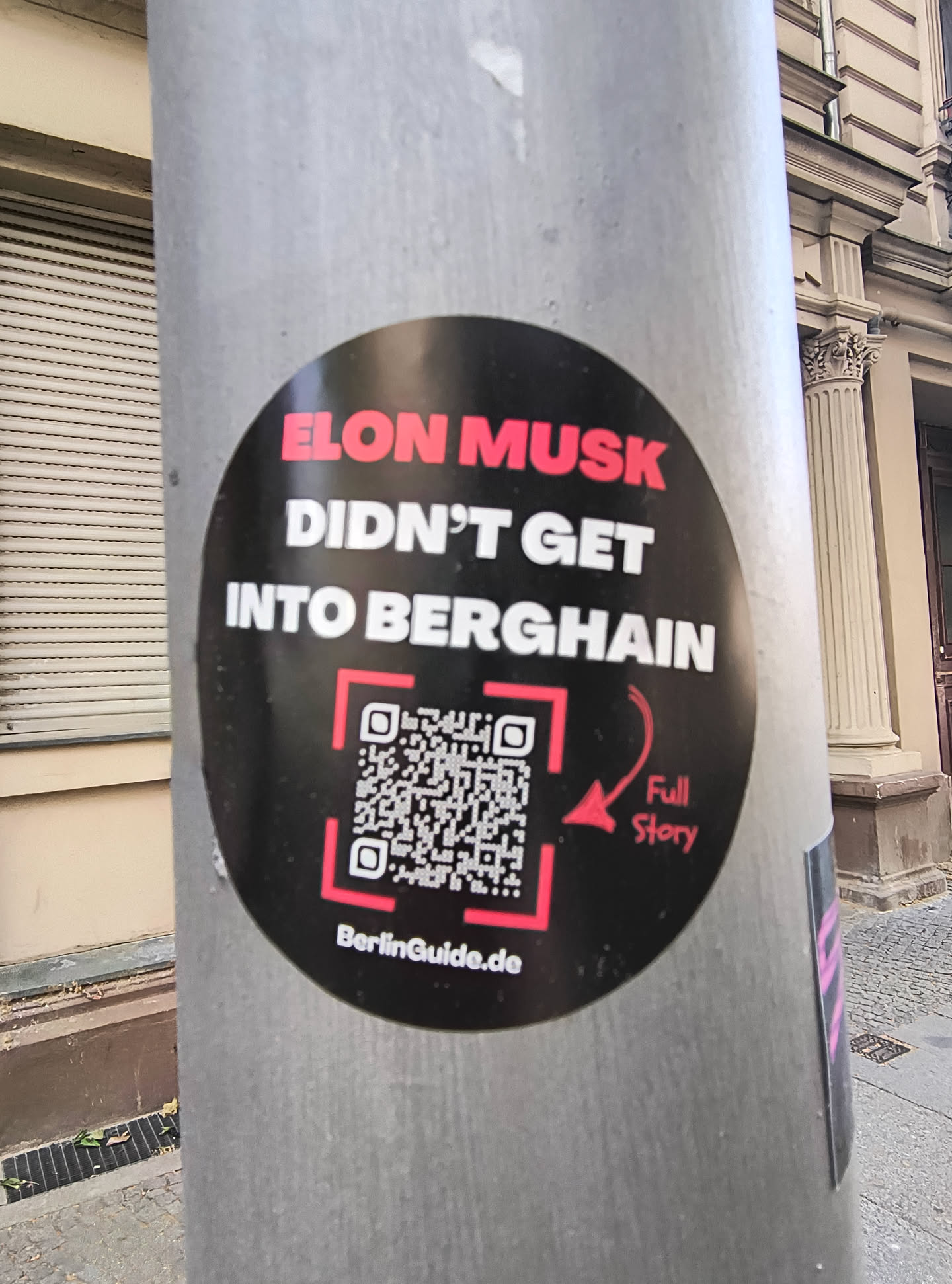 This is a round black sticker attached to a metal pole. The text says, in all caps, "Elon Musk DIDN'T GET INTO BERGHAIN" with a QR code and the text "Full Story" next to it, as well as "BerlinGuide.de".