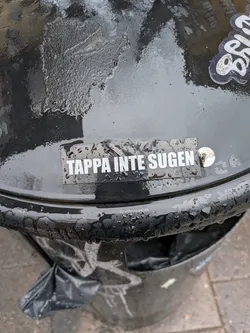 The sticker is rectangular with a black background and features white, uppercase sans-serif text. It reads 'TAPPA INTE SUGEN'. The sticker is applied to a dark, wet, possibly metallic surface, likely a trash can, with numerous water droplets visible on and around it, giving it a slightly reflective and textured appearance. The design is minimalist and text-focused.