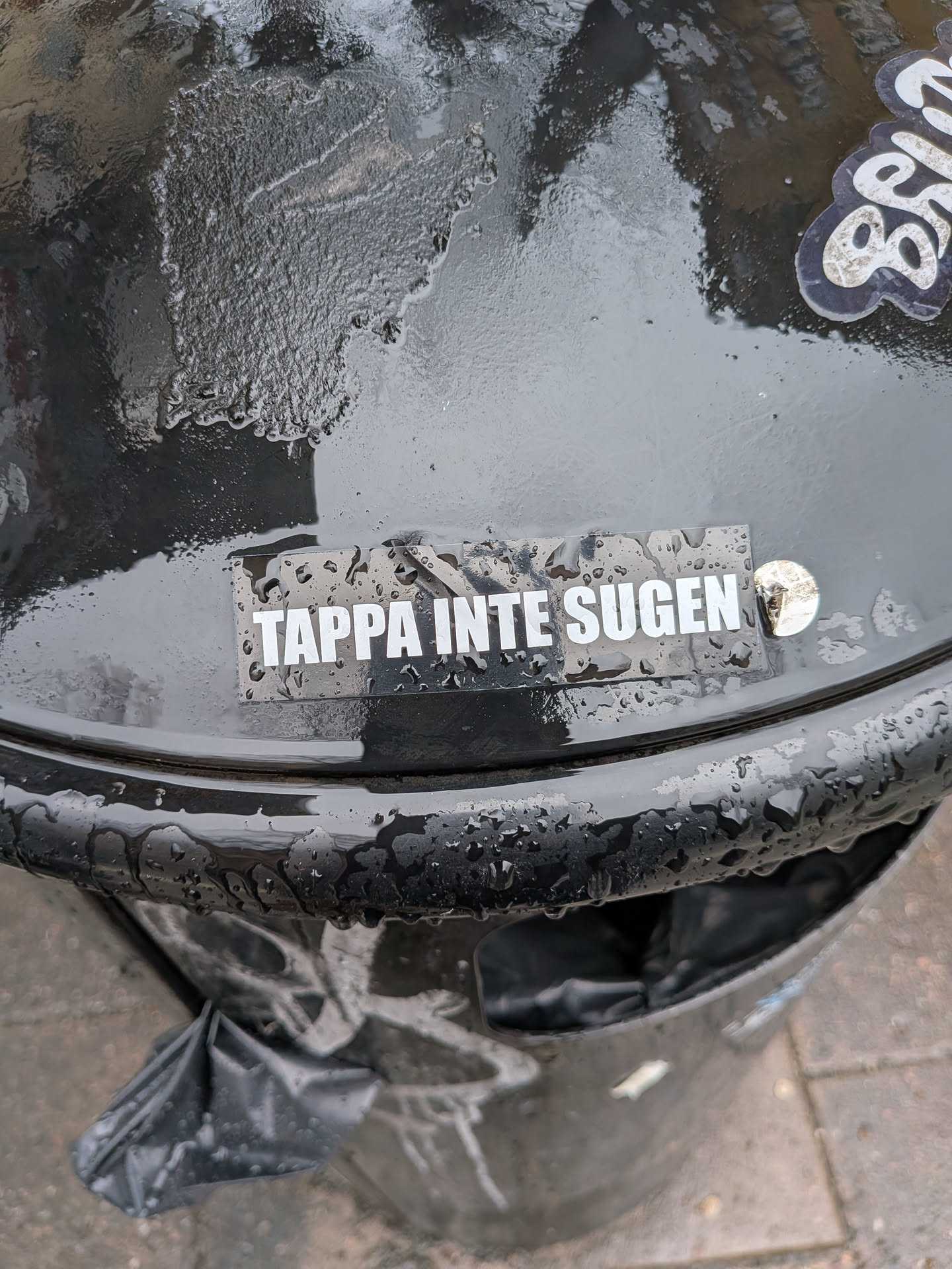 The sticker is rectangular with a black background and features white, uppercase sans-serif text. It reads 'TAPPA INTE SUGEN'. The sticker is applied to a dark, wet, possibly metallic surface, likely a trash can, with numerous water droplets visible on and around it, giving it a slightly reflective and textured appearance. The design is minimalist and text-focused.
