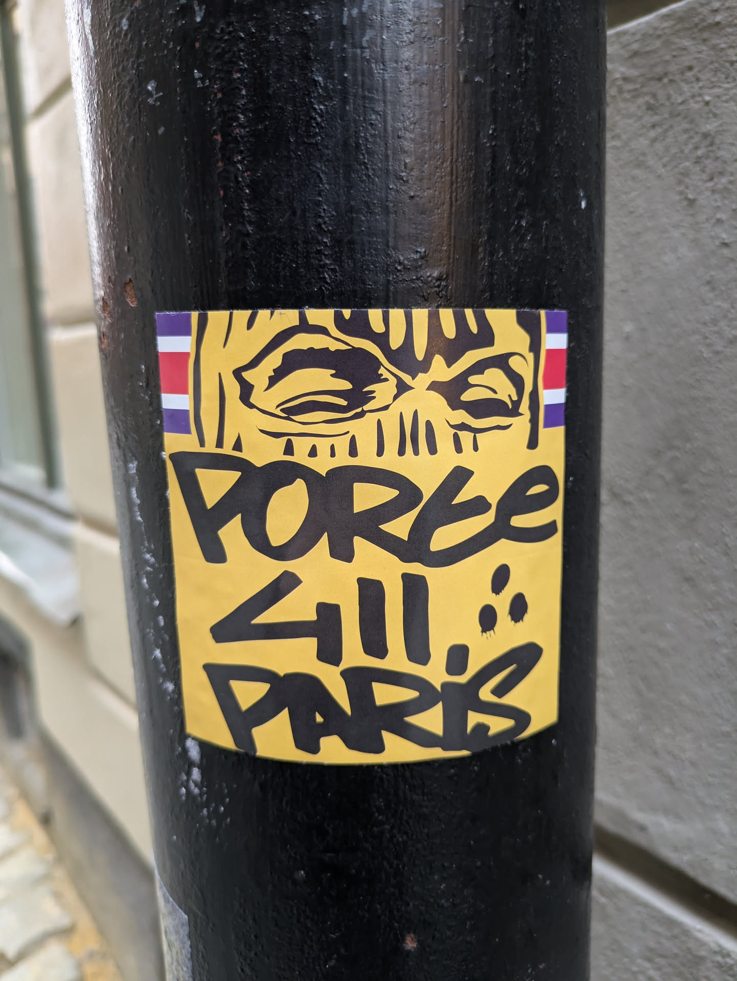 Yellow sticker with black graffiti art of a face wearing a balaclava.  The text 'POREE 411 PARIS' is written below the image. A small flag is visible in the upper left corner of the sticker.