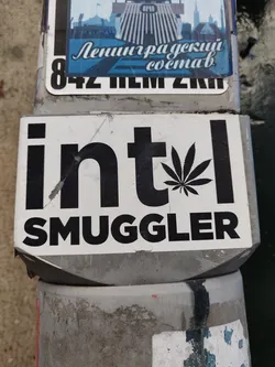 A sticker with the text "intel smuggler" and a cannabis leaf. The sticker is black and white and is placed on a grey surface.
