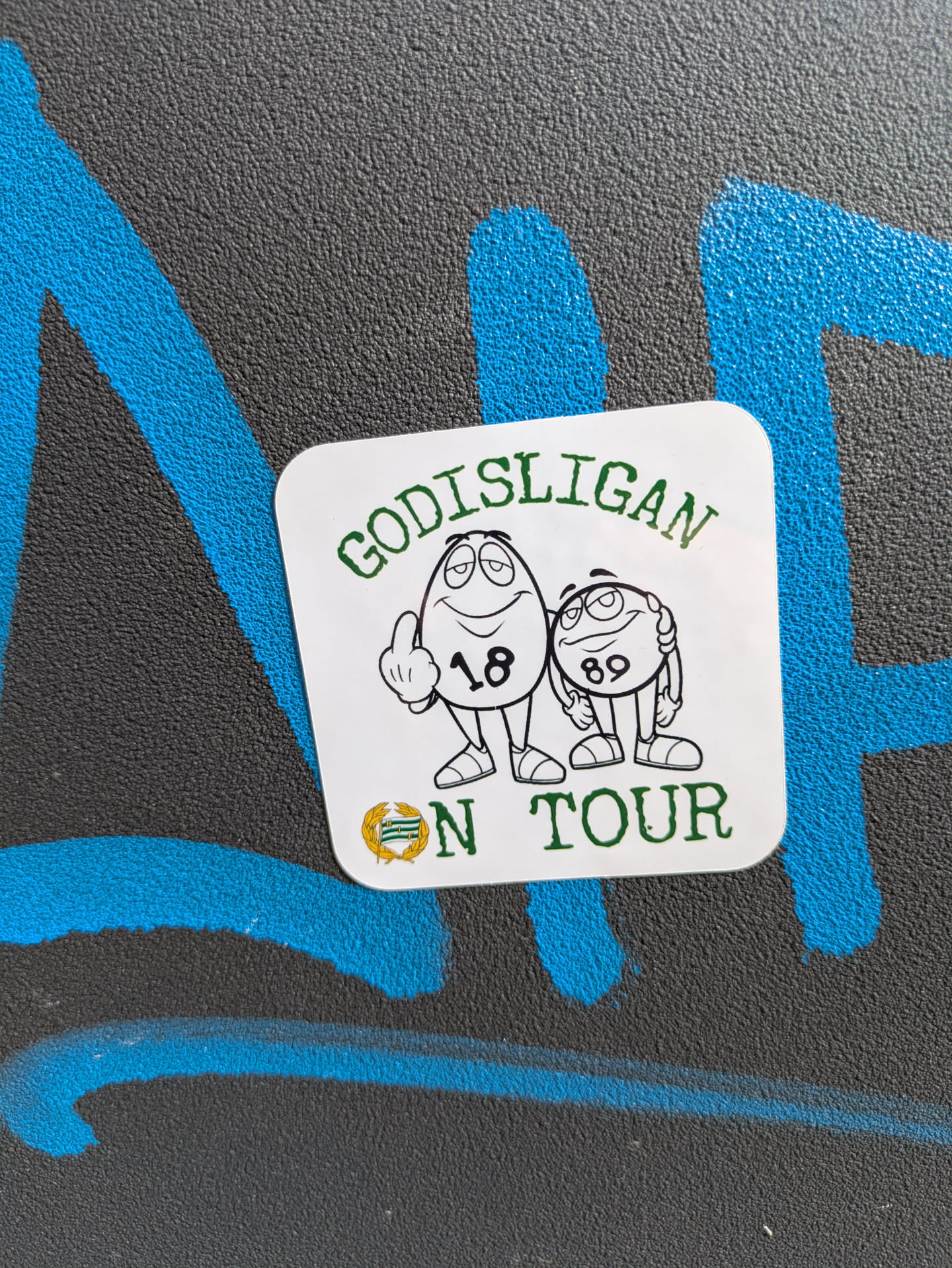 A square sticker features two cartoon characters reminiscent of M&M candies. The characters are outlined in black with green details. The first character displays '18' on its body and is gesturing with its middle finger raised. The second character shows '89' on its body. Above the characters, the text 'GODISLIGAN' is printed in green. Below, the words 'ON TOUR' are also written in green, flanked by a green and gold emblem.