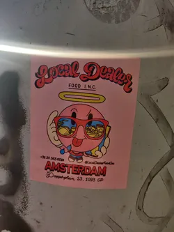 Pink sticker with an illustration of a cartoon character wearing sunglasses. It has the name and address of a food establishment in Amsterdam.