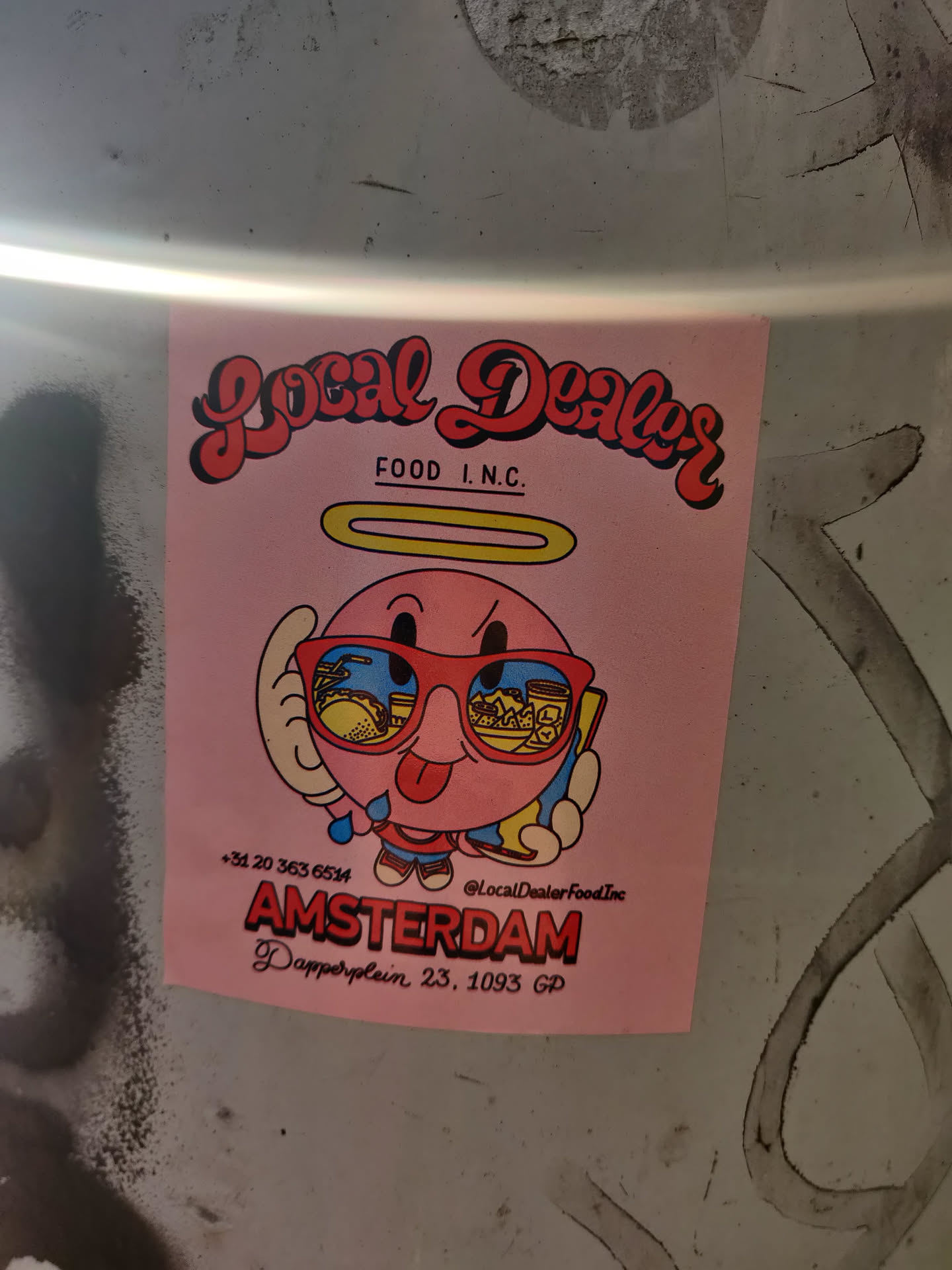 Pink sticker with an illustration of a cartoon character wearing sunglasses. It has the name and address of a food establishment in Amsterdam.