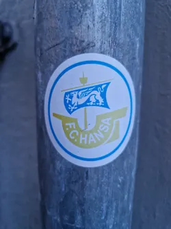 A circular sticker featuring the logo of F.C. Hansa Rostock, a German football club. The logo depicts a griffin on a ship's sail within a circular frame. The sticker is affixed to a weathered metal pipe.