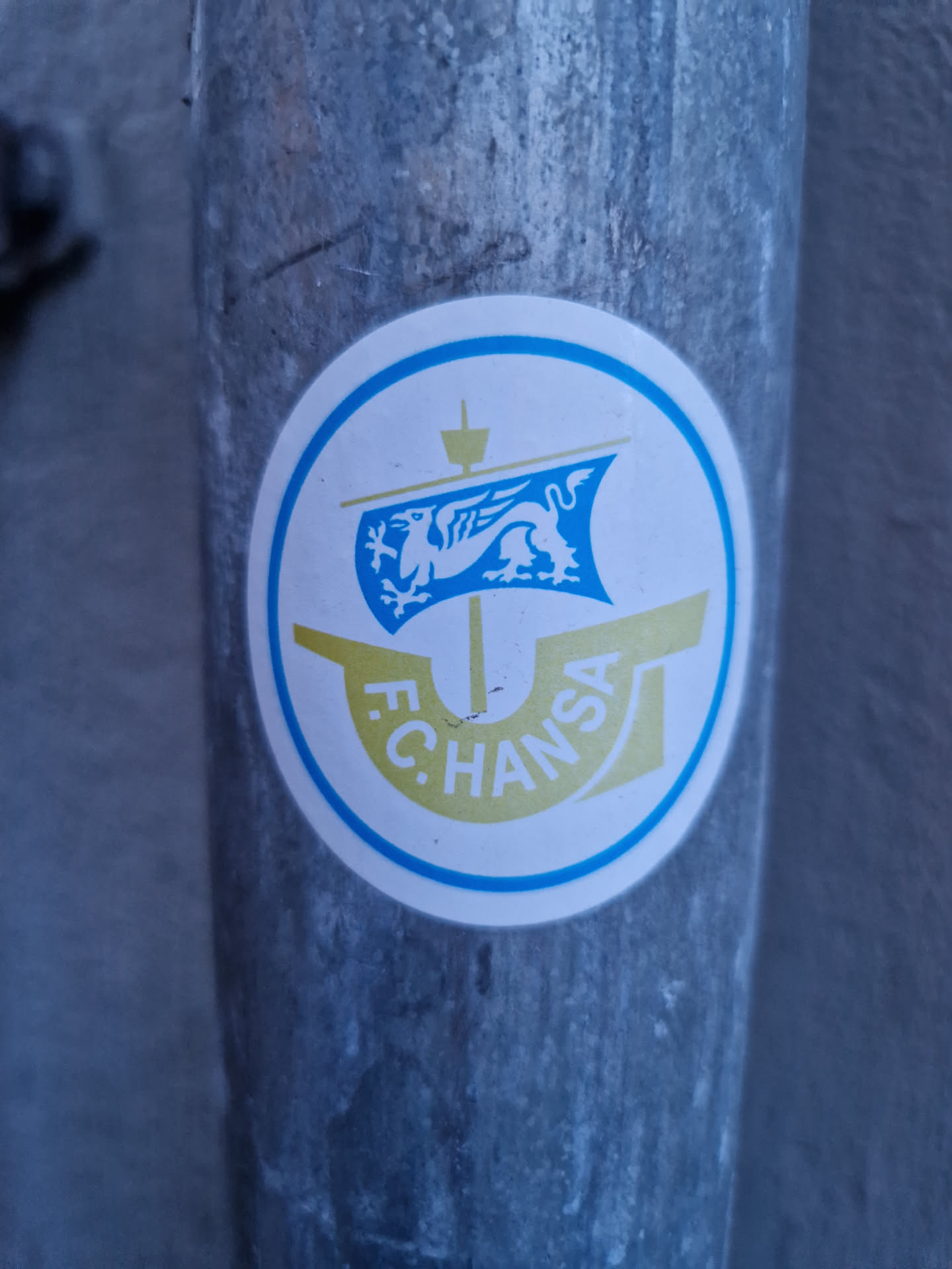 A circular sticker featuring the logo of F.C. Hansa Rostock, a German football club. The logo depicts a griffin on a ship's sail within a circular frame. The sticker is affixed to a weathered metal pipe.