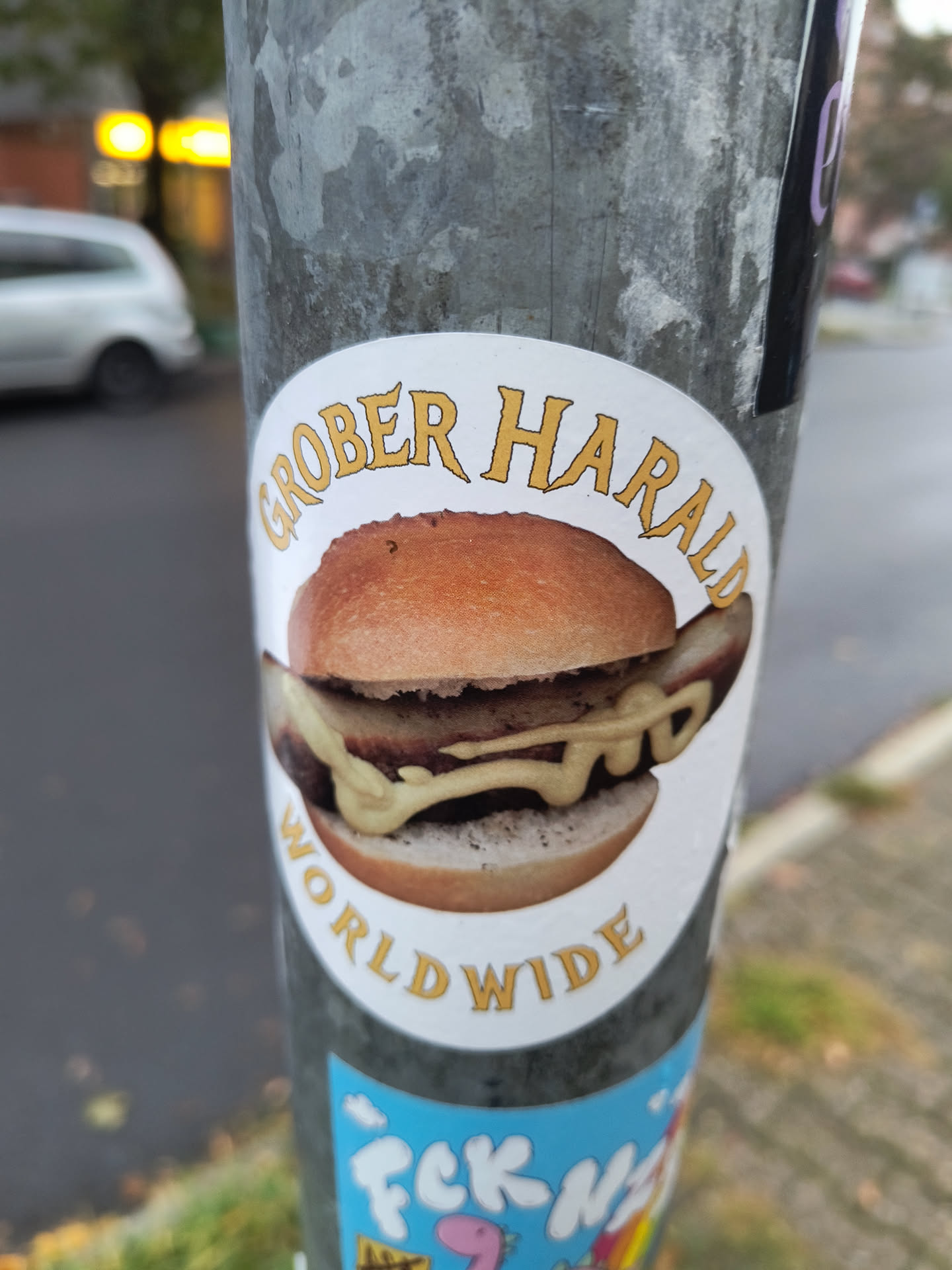 The sticker features a realistic depiction of a hamburger with a sausage patty and generous amounts of mustard. The bun is golden brown, and the sausage appears to be grilled. The text 'GROBER HARALD' is arched above the burger in a stylized, yellow font with a darker outline. Below the burger, also in a similar yellow font, is the word 'WORLDWIDE', also arched. The sticker has a white background with a thin white border.