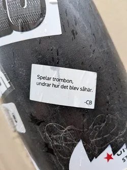 A rectangular white sticker with slightly rounded corners, featuring black sans-serif text. The sticker is applied to a dark, textured surface, possibly metal or plastic, and has several water droplets on its surface, indicating exposure to moisture. The text is centered on the sticker.