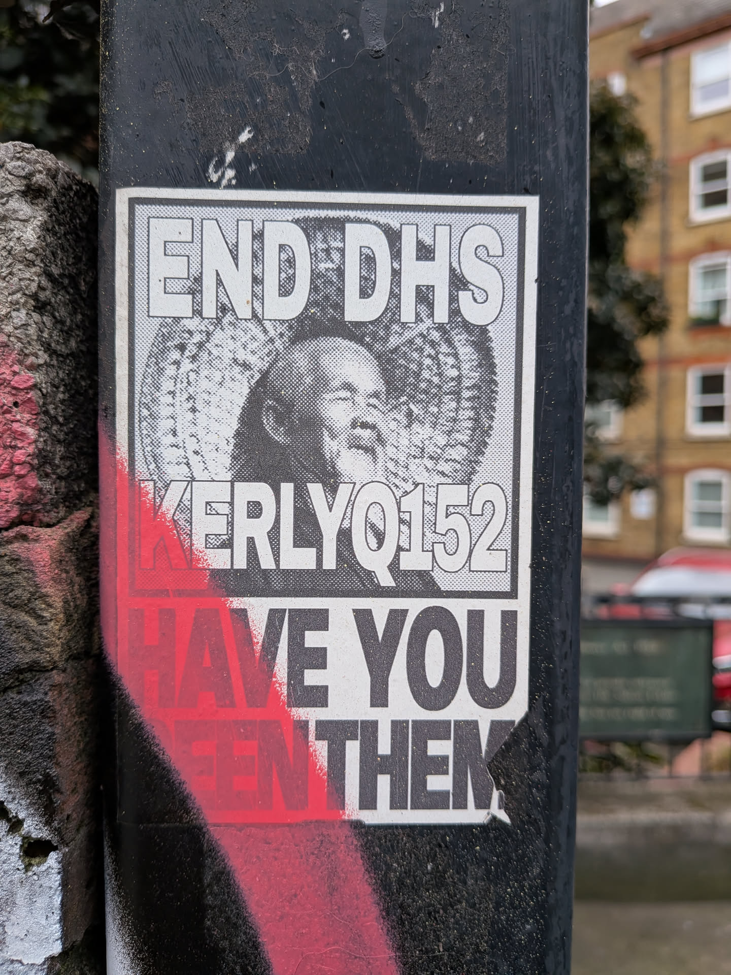 A rectangular, black and white sticker affixed to a dark, textured pole. The sticker features a halftone portrait of an older man with a beard, smiling, centered within a circular, radiating pattern. Above the portrait, in bold, sans-serif capital letters, is the text 'END DHS'. Below the portrait, also in bold capital letters, is 'KERLYQ152'. The bottom section of the sticker reads 'HAVE YOU SEEN THEM', but the words 'HAVE YOU SEEN' are heavily obscured by vibrant red spray paint that streaks across the lower left portion of the sticker and the pole. The overall aesthetic is stark and impactful, with a clear protest or activist message. The sticker shows some minor wear and tear, particularly a slight tear at the bottom right corner.