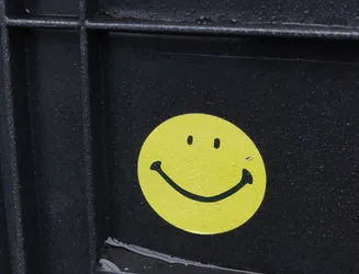 A yellow smiley face sticker is affixed to a dark gray surface.  The sticker appears to be vinyl and has a simple design. Water droplets are visible on the sticker and the surrounding surface.