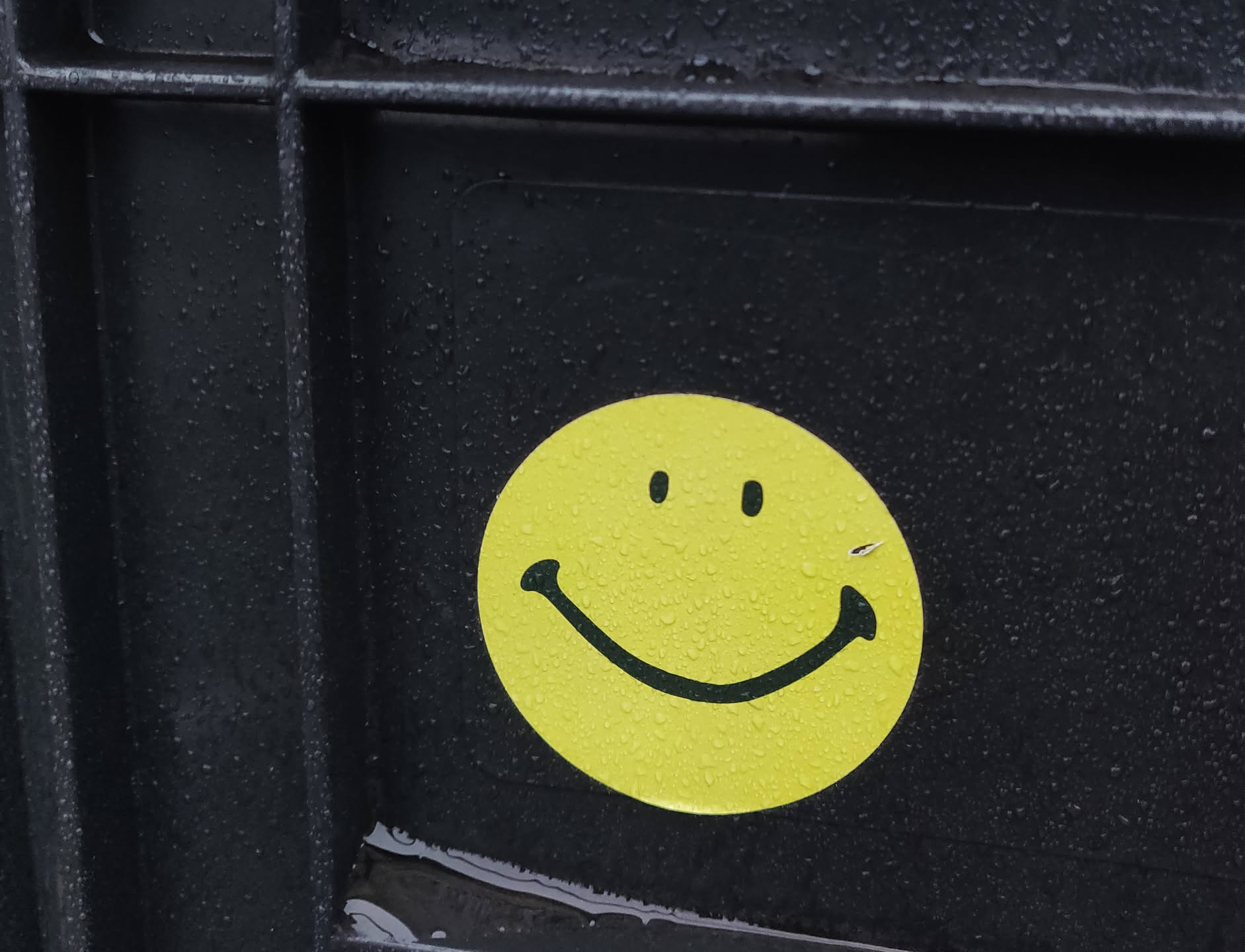 A yellow smiley face sticker is affixed to a dark gray surface.  The sticker appears to be vinyl and has a simple design. Water droplets are visible on the sticker and the surrounding surface.