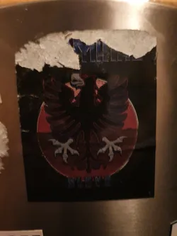 A sticker featuring a stylized Albanian eagle emblem on a dark background. The eagle is positioned in a circular design with a red gradient. The text "SLIVE" is visible at the bottom. The sticker appears to be slightly damaged around the edges.