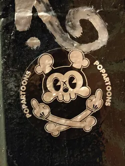Round black and white sticker with a skull character in a pop art style. The skull has big ears and is showing the middle finger with both hands. Crossed bones are behind the skull. The text "POPARTOONS" is written around the sticker.