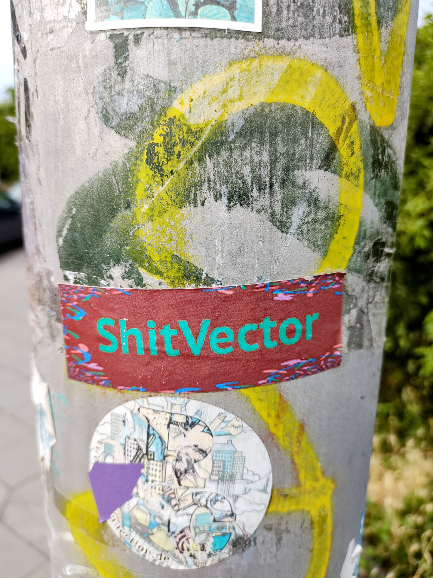 A rectangular sticker with the text "ShitVector" written in teal on a maroon background. The sticker has a slightly worn look and is adhered to a gray pole alongside other stickers and graffiti.