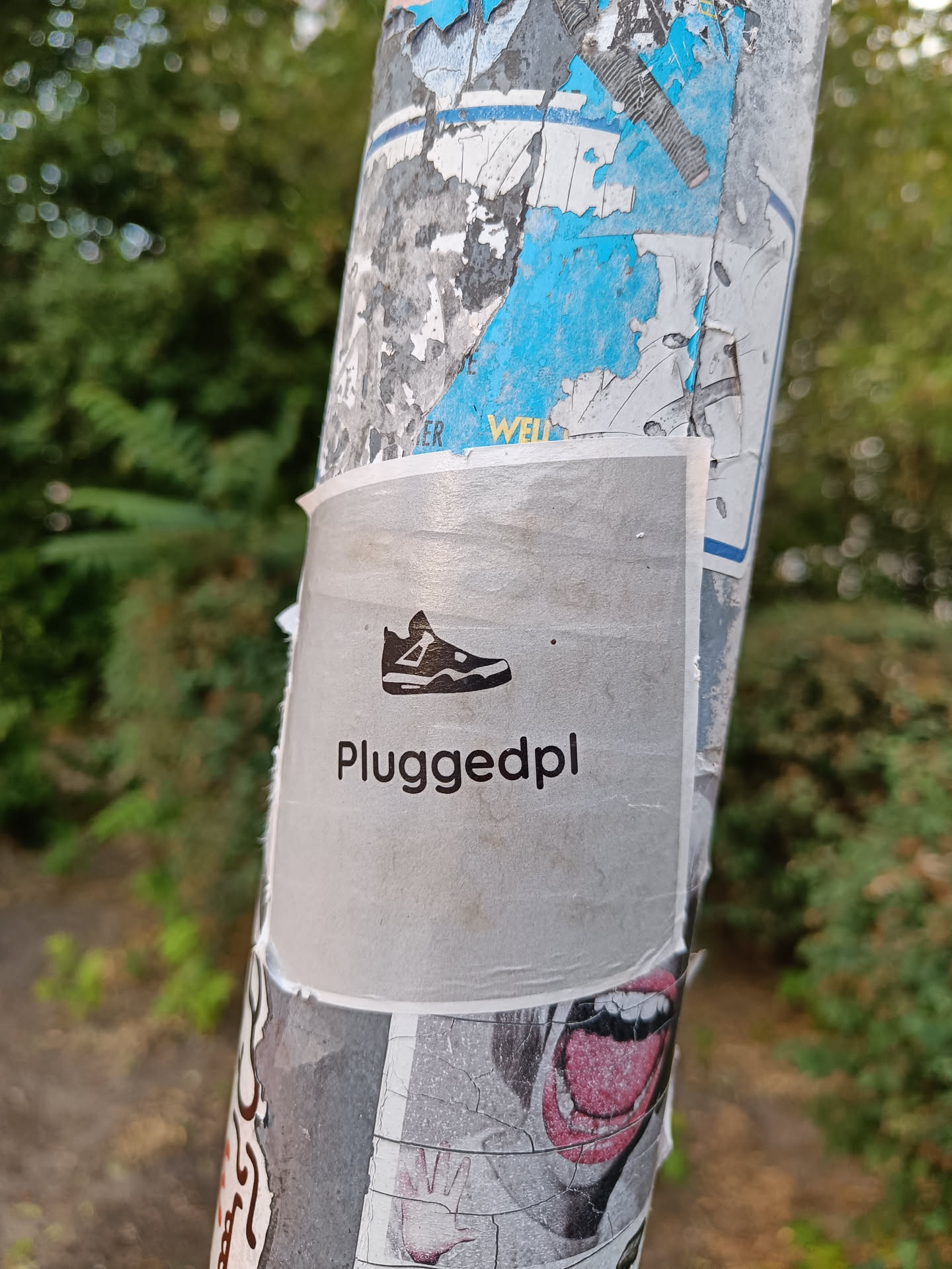 A silver sticker with a black silhouette of a sneaker and the text "Pluggedpl" in a sans-serif font. The sticker is affixed to a pole covered in other stickers and graffiti.