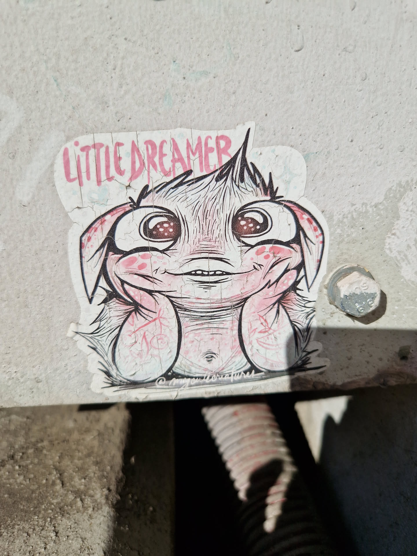 A sticker featuring a cute cartoon creature with big eyes and rosy cheeks. The creature is sitting with its hands on its cheeks, and the words "Little Dreamer" are written above it. The sticker has a slightly worn appearance.