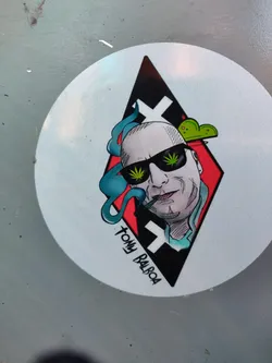Round sticker featuring an illustration of Tommy Balboa, a bald man wearing sunglasses, smoking a joint. The illustration is in a diamond shape with a red and black background. The style is reminiscent of tattoo art.