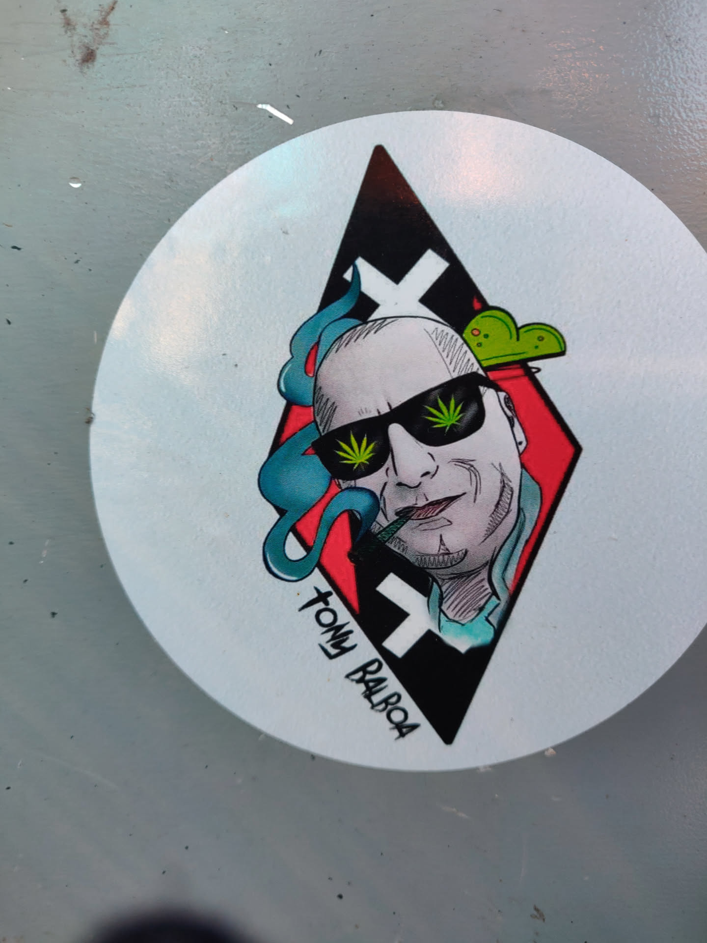 Round sticker featuring an illustration of Tommy Balboa, a bald man wearing sunglasses, smoking a joint. The illustration is in a diamond shape with a red and black background. The style is reminiscent of tattoo art.