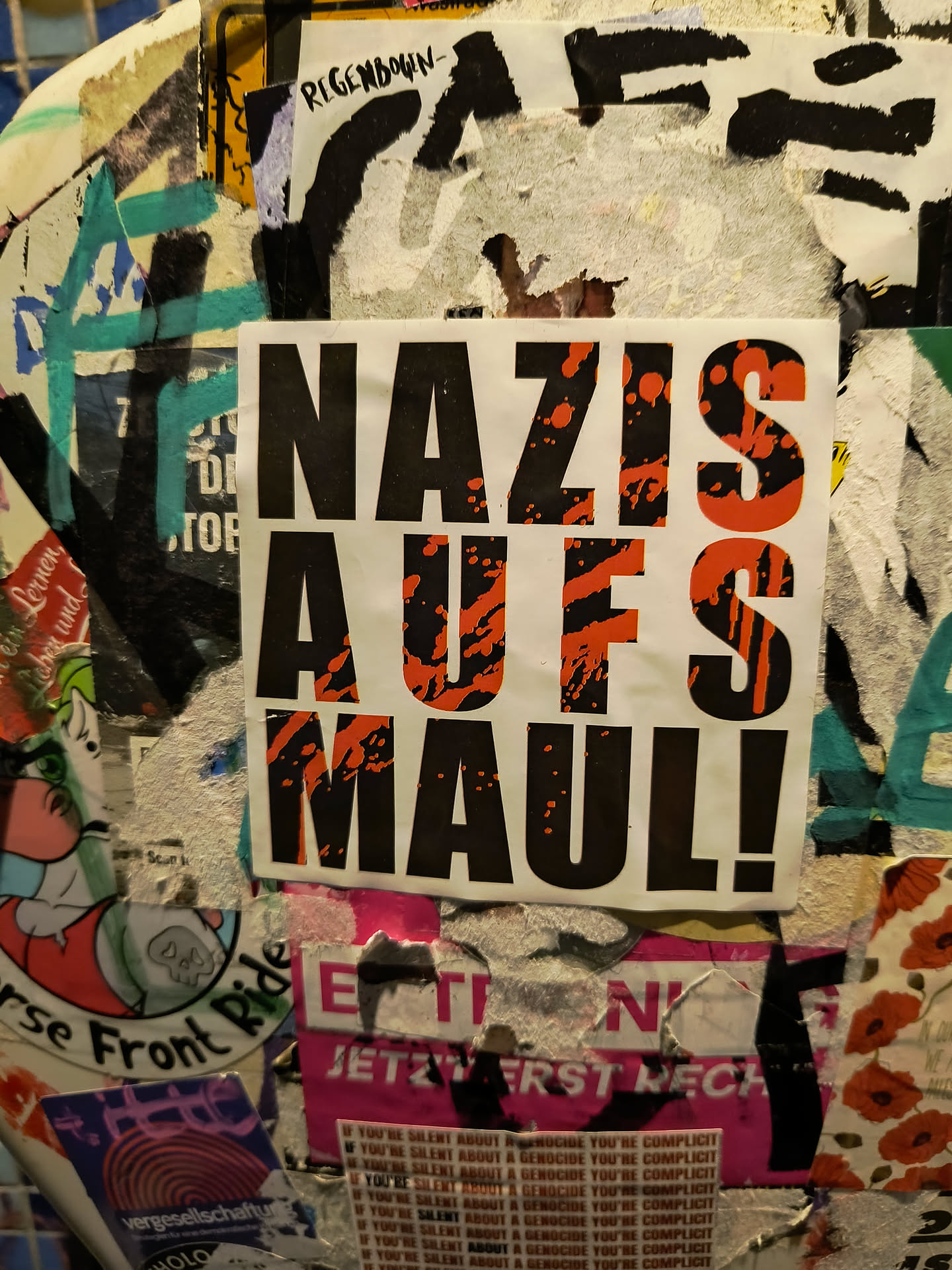 A sticker with the text "Nazis aufs Maul!" in black and orange. The background is white. The text is bold and in all caps. The sticker is attached to a surface with other stickers.