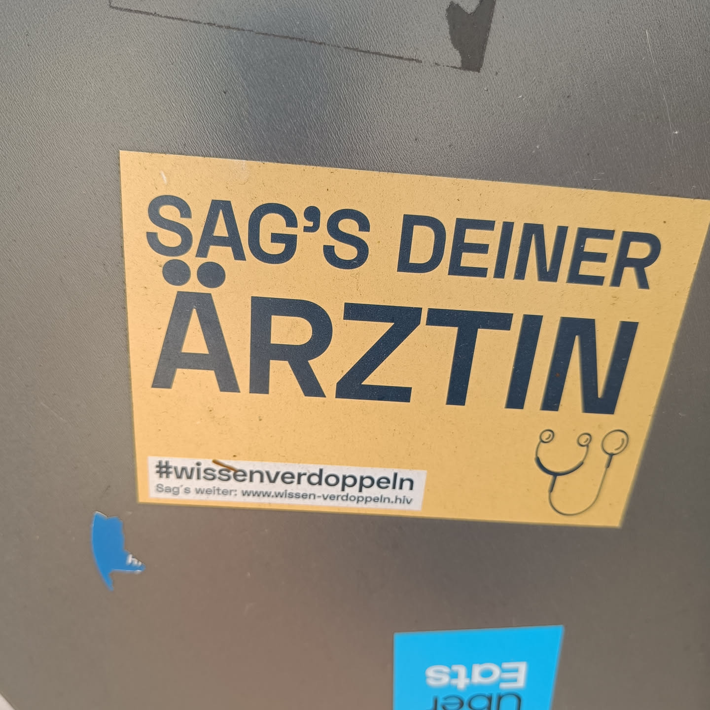 Yellow sticker with the text "Sag's deiner Ärztin" (Tell your doctor) and a small drawing of a stethoscope.  It also includes a hashtag and website address at the bottom.