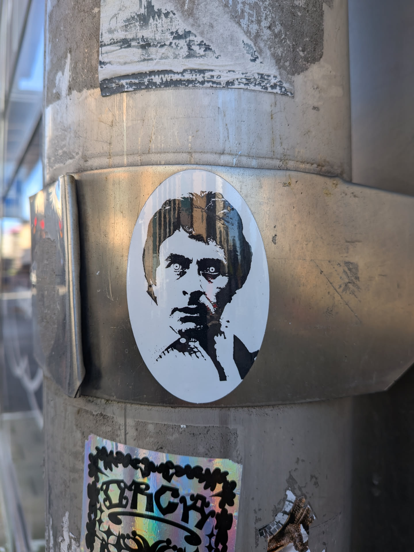An oval-shaped vinyl sticker displaying a black-and-white, high-contrast stencil-style portrait of actor Gene Wilder, depicted with an intense, wide-eyed stare and wavy hair reminiscent of his iconic role in 'Young Frankenstein'. The artwork relies heavily on stark negative space and dark shadows. It is adhered to a brushed metal band wrapped around an outdoor pole.