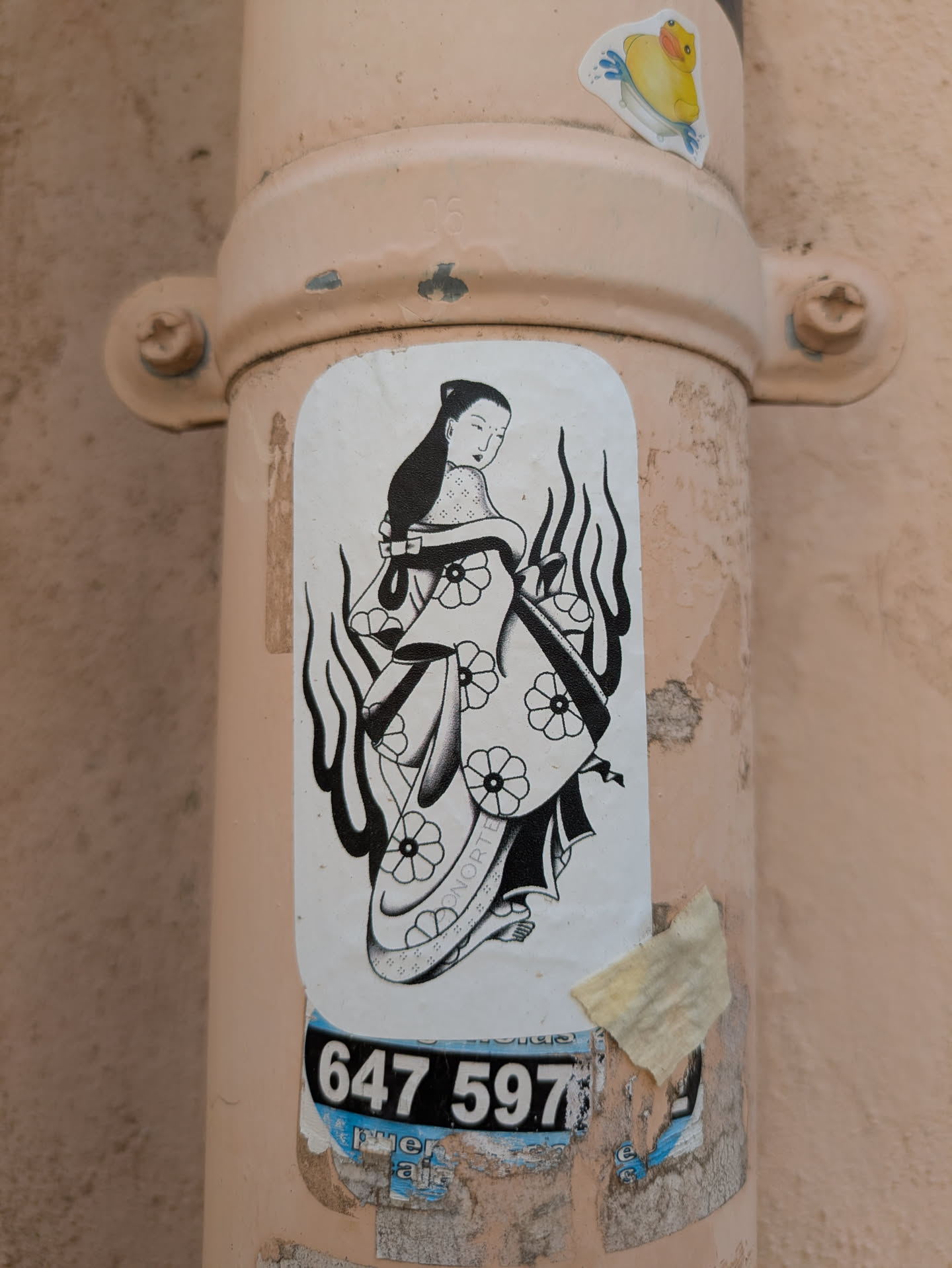 The sticker depicts a black and white geisha figure adorned with floral patterns on her kimono, surrounded by stylized flames. It is stuck on a peach colored pipe, near a number and another sticker of a rubber duck.