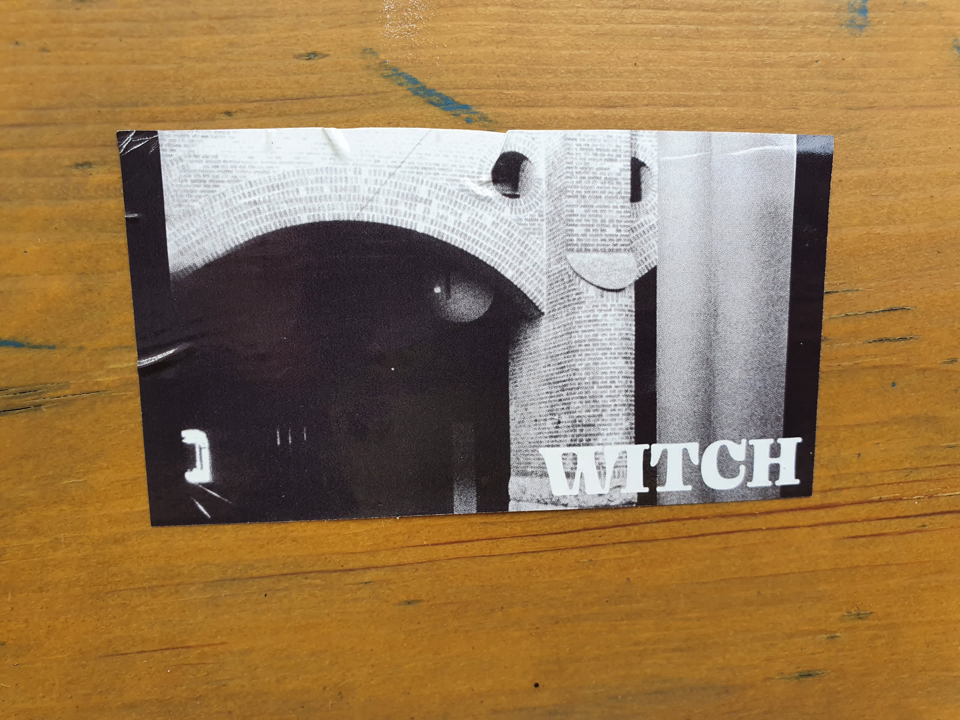 A black and white photo sticker featuring an archway made of bricks. The word "WITCH" is printed in white sans-serif font at the bottom right. There's a mysterious, almost eye-like shape visible in the dark archway. The sticker has a slightly worn appearance, suggesting it has been placed outdoors.