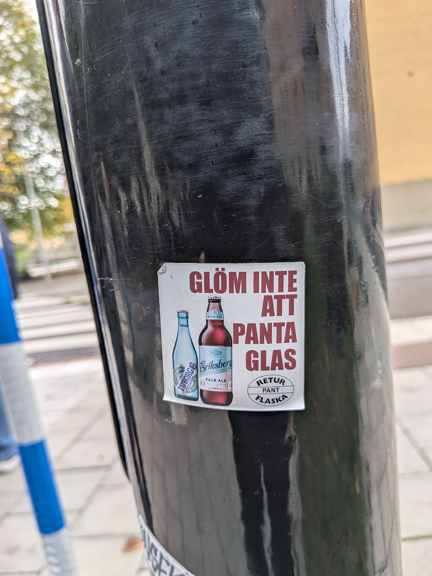 A square sticker with a white background, affixed to a black metal pole. It features bold red-brown Swedish text that reads 'GLÖM INTE ATT PANTA GLAS'. To the left of the text are illustrations of two beer bottles: a light blue Tuborg bottle and a brown Eriksberg Pale Ale bottle. In the bottom right corner, there is a circular logo containing the text 'RETUR PANT FLASKA'.