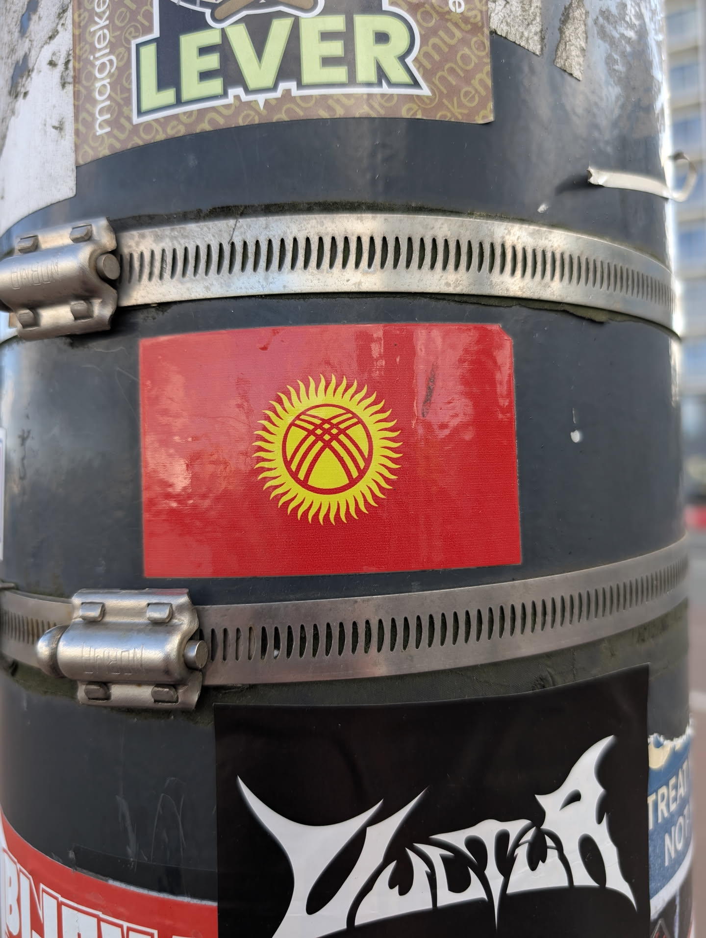 The sticker is rectangular and displays the flag of Kyrgyzstan. The background of the flag is red, symbolizing bravery and wealth. In the center, there is a yellow sun with 40 rays, representing the unity of 40 Kyrgyz tribes. Within the sun, there is a depiction of a tunduk, the crown of a yurt, stylized with crossed lines, symbolizing the traditional dwelling and the universe.