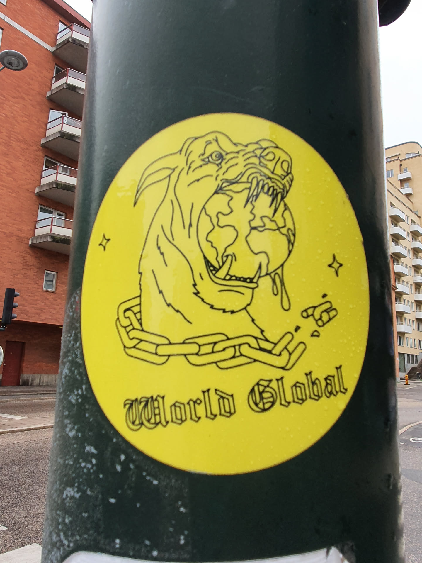 A yellow round sticker with a drawing of a wolf or dog holding the Earth in its mouth. The animal is chained. The text "World Global" is written below the drawing. The background is yellow with some small details.