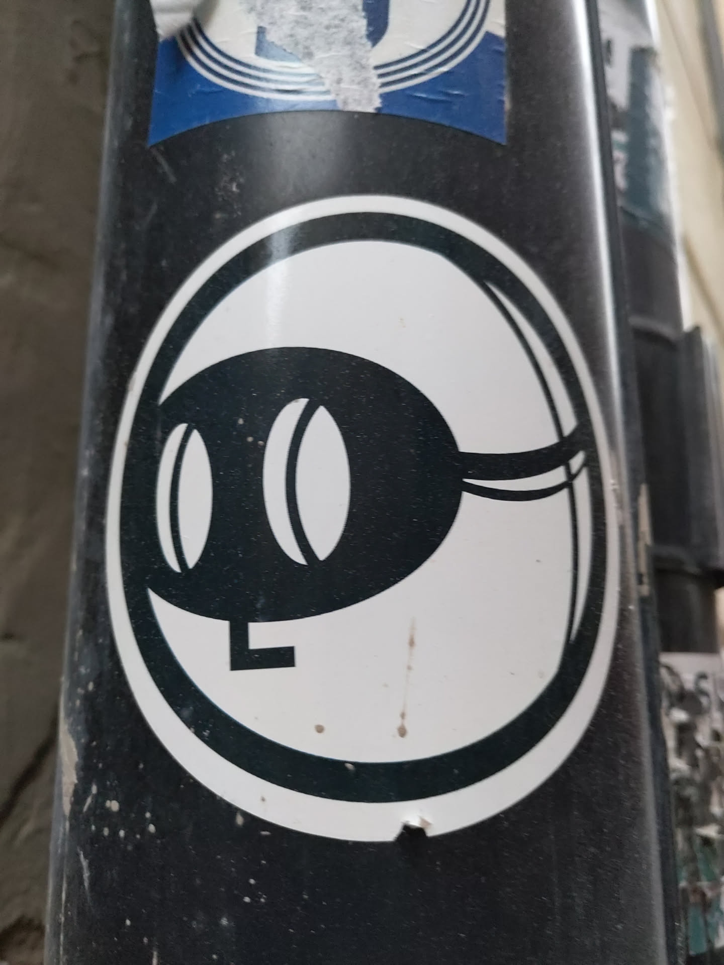 A black and white circular sticker featuring a simple, abstract design. The main element seems to be a stylized creature or object with two large, oval eyes and a curved body. The style is clean and graphic. The sticker appears to be affixed to a dark-colored pole or surface.