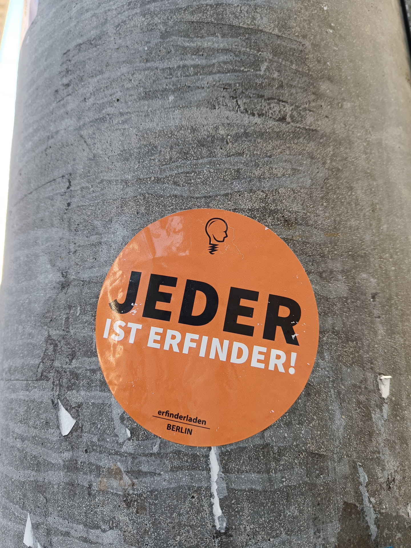 An orange round sticker with the text "Jeder ist Erfinder!" in black bold letters. At the bottom, it says "erfinderladen BERLIN" in a smaller font.  A small logo depicting a light bulb with a head is present at the top. The sticker is affixed to a grey concrete surface.