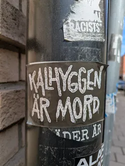 A sticker with the text 'Kalhyggen är mord' is stuck on a black pole. The text is in white with a dripping effect, set against a black background. The sticker appears to be partially torn at the edges, suggesting it has been there for some time. The pole shows signs of wear and tear, with other stickers and residue from past postings visible. The overall aesthetic is reminiscent of urban environments where such forms of expression are common.