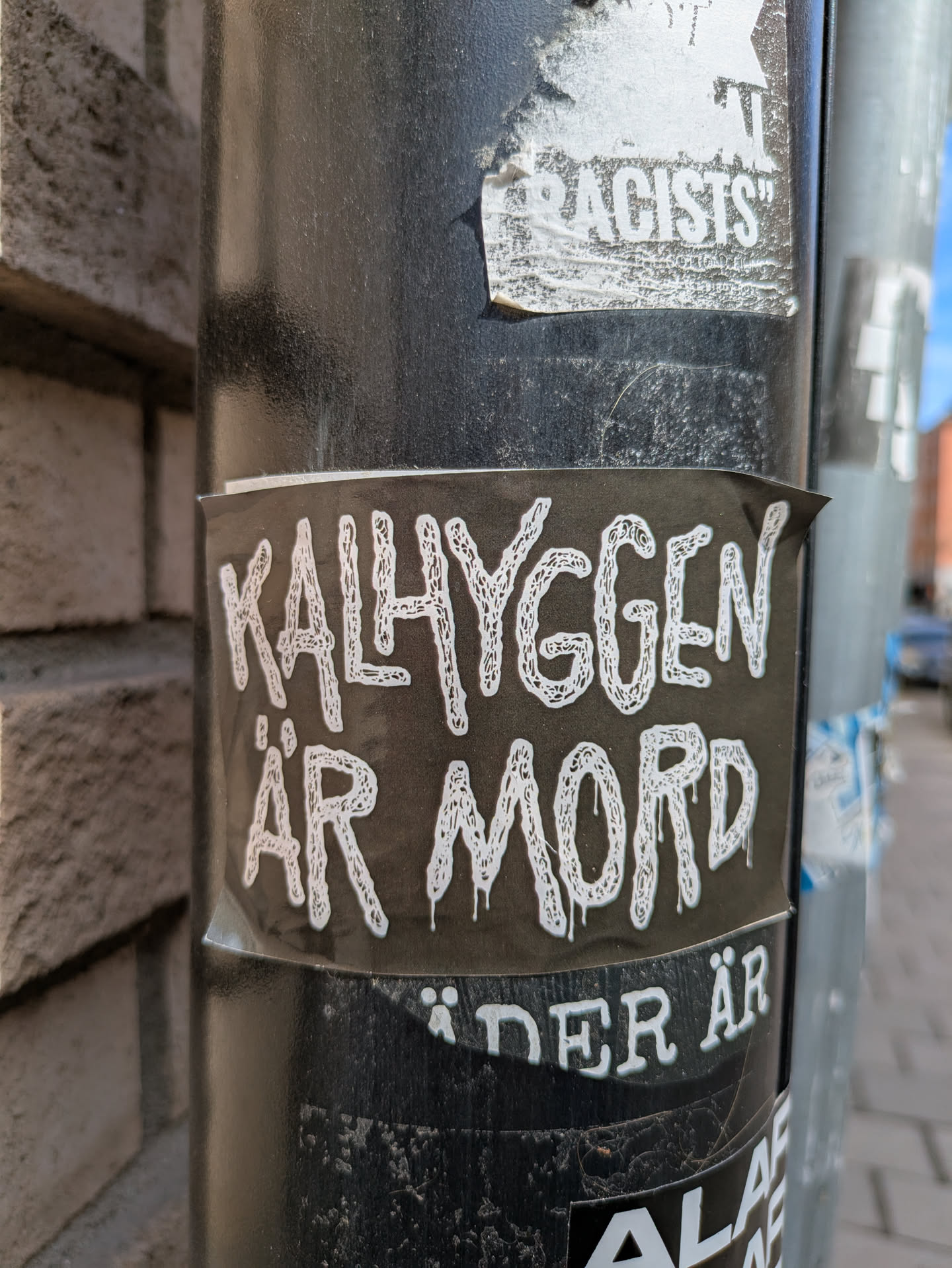A sticker with the text 'Kalhyggen är mord' is stuck on a black pole. The text is in white with a dripping effect, set against a black background. The sticker appears to be partially torn at the edges, suggesting it has been there for some time. The pole shows signs of wear and tear, with other stickers and residue from past postings visible. The overall aesthetic is reminiscent of urban environments where such forms of expression are common.