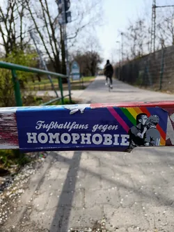 A sticker placed on a barrier shows two men kissing with a rainbow flag in the background. The text on the sticker says "Fußballfans gegen Homophobie" which translates to "Football fans against homophobia".