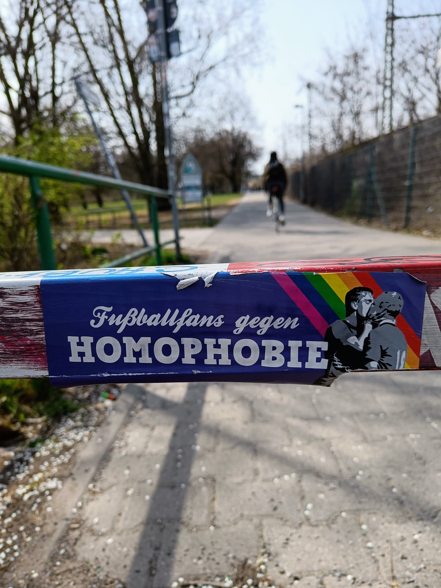 A sticker placed on a barrier shows two men kissing with a rainbow flag in the background. The text on the sticker says "Fußballfans gegen Homophobie" which translates to "Football fans against homophobia".