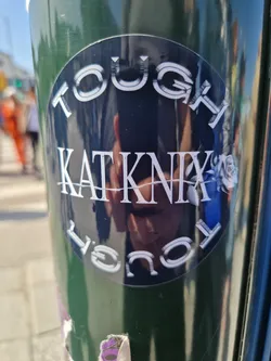A round black and white sticker with the text "Tough Kat Knix" in a stylized font. There is a small rose design next to the name. The sticker is placed on a green pole.