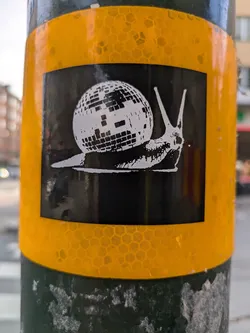 A black and white sticker of a snail carrying a disco ball on a yellow reflective background. The style is simple and graphic.