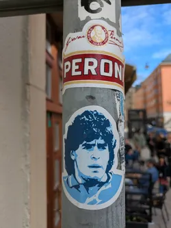 The sticker features a blue and white portrait of Diego Maradona. It is placed on a pole with other stickers. The portrait is stylized, with shades of blue and white creating a contrast. The pole is textured with signs of wear and other stickers. There's a Peroni sticker above it.