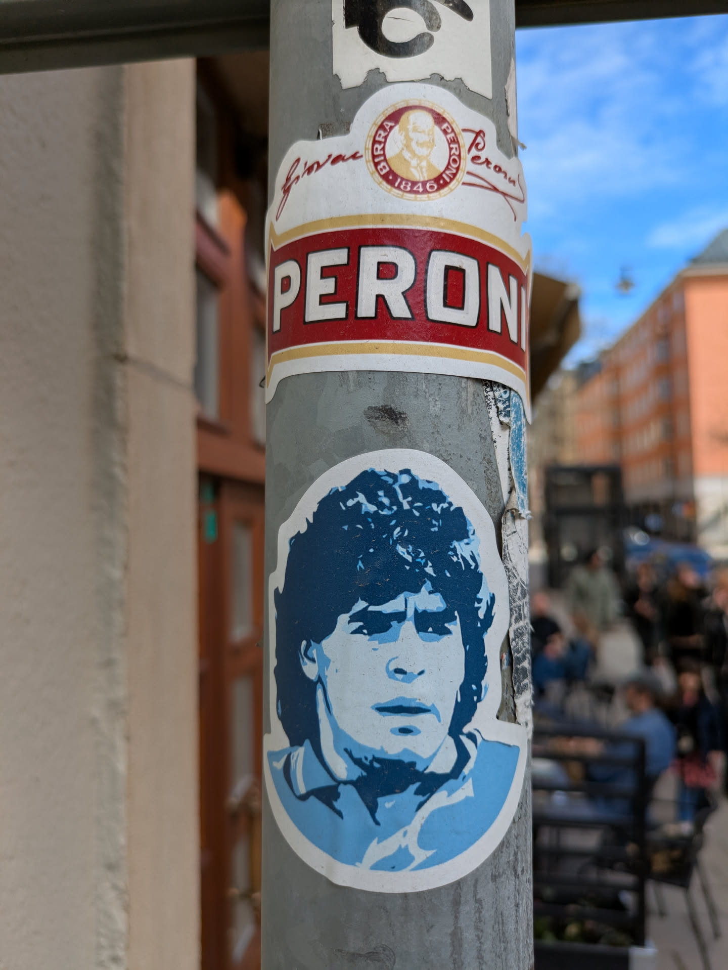 The sticker features a blue and white portrait of Diego Maradona. It is placed on a pole with other stickers. The portrait is stylized, with shades of blue and white creating a contrast. The pole is textured with signs of wear and other stickers. There's a Peroni sticker above it.