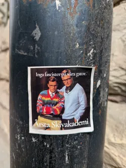 This is a sticker featuring two men. The man on the left is wearing a colorful sweater with a tie, and the man on the right is wearing a light blue sweater. The text above them reads "Inga fascister på våra gator", which translates to "No fascists in our streets". Below the image, it says "Årsta Skivakademi".