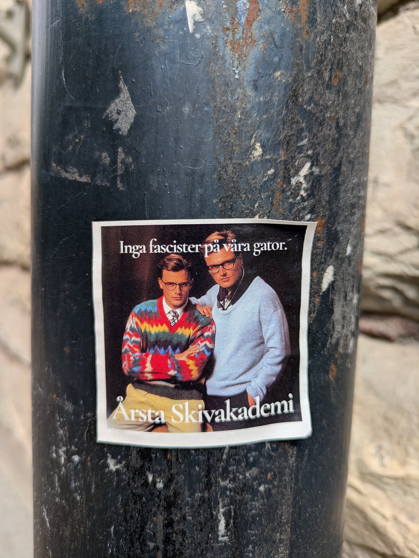 This is a sticker featuring two men. The man on the left is wearing a colorful sweater with a tie, and the man on the right is wearing a light blue sweater. The text above them reads "Inga fascister på våra gator", which translates to "No fascists in our streets". Below the image, it says "Årsta Skivakademi".