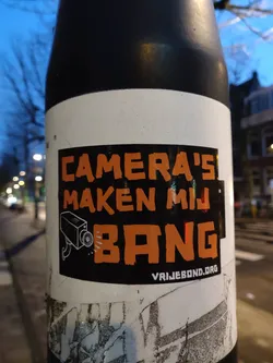 A sticker expressing unease about surveillance cameras. It features a stylized depiction of a security camera and the text "CAMERA'S MAKEN MIJ BANG" which translates from Dutch to English as "Cameras make me nervous".  The sticker also includes a website address.