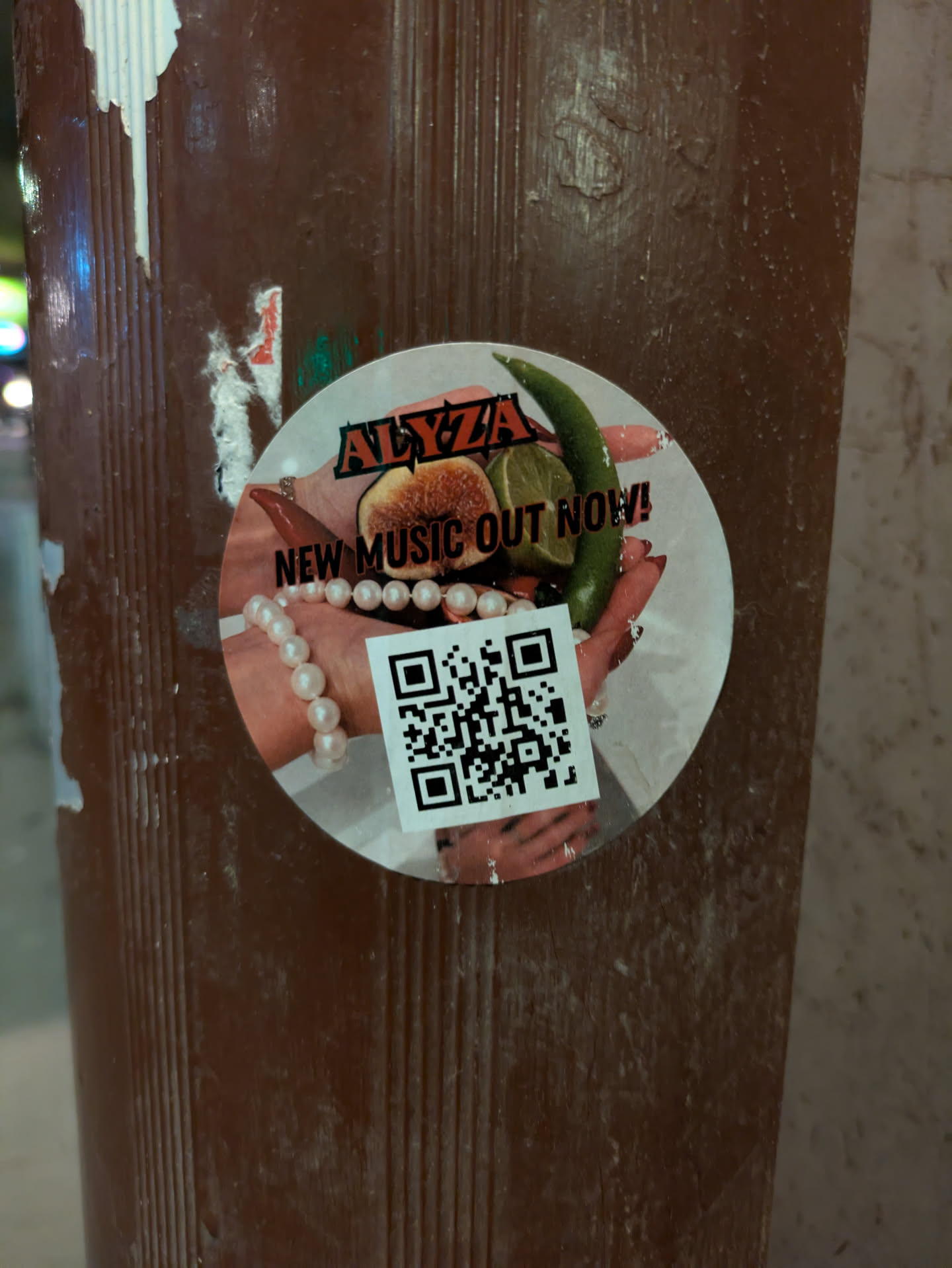This round sticker is promoting new music from Alyza. It features a pair of hands holding fruit and a green pepper, adorned with rings and a pearl bracelet.  The sticker also includes a QR code.