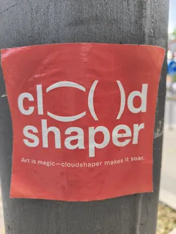 A red sticker on a gray light pole with the words \"cloud shaper\" written on it in white. The logo uses parenthesis in place of the \"o\" and the tagline reads \"Art is magic - cloudshaper makes it soar.\"