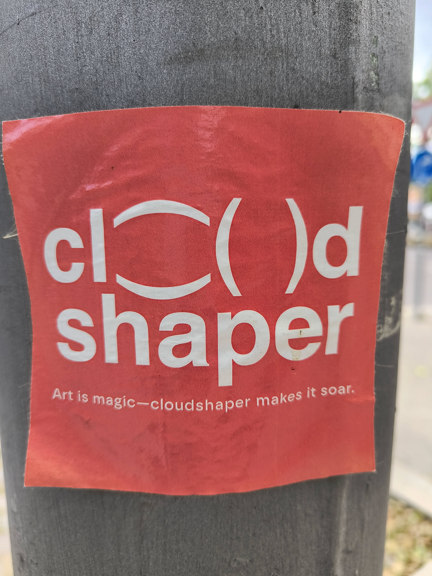 A red sticker on a gray light pole with the words \"cloud shaper\" written on it in white. The logo uses parenthesis in place of the \"o\" and the tagline reads \"Art is magic - cloudshaper makes it soar.\"
