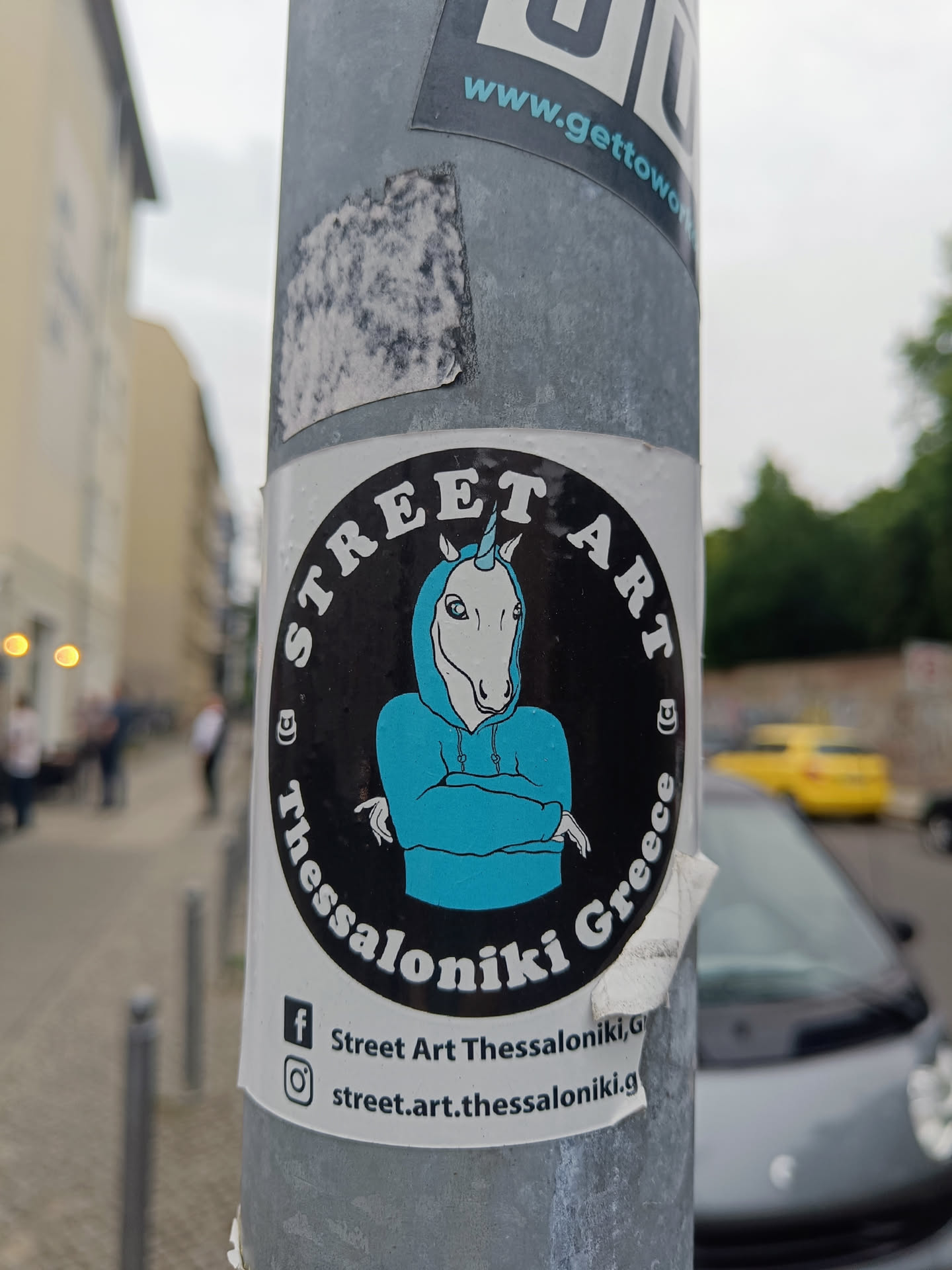 Round sticker featuring a unicorn wearing a blue hoodie with its arms crossed. The text 'Street Art' is at the top, with 'Thessaloniki Greece' at the bottom.  Social media links for Facebook and Instagram are at the bottom.